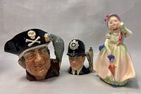 Three ceramic pieces side-by-side: pirate character mug, British policeman character mug, and Royal Doulton girl figurine