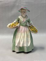 Front view of the Royal Doulton figurine showing the woman in a green dress and pink apron holding baskets of yellow flowers.