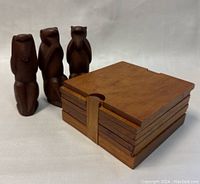 Set of six square wooden coasters stacked in a wooden holder next to three carved wooden wise monkey figurines.