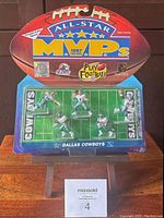 Overall front view of the sealed 1997 All Star MVPs Dallas Cowboys figurine set showing 5 player miniatures on a football field base inside original display packaging shaped like a football.