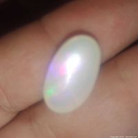 Close-up image of the opal gemstone showing its translucent body with play-of-color in purple, blue, green, red, and orange hues. The stone is held between fingers, highlighting its size and surface.