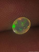 Close-up photo of small oval Australian rainbow fire opal showing vibrant green and orange internal color flashes on finger background.