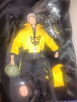 Photo of G.I. Joe 12-inch figure lying down wearing yellow jacket and black pants with yellow stripes, with camouflage helmet, black chest harness, yellow cloth item, and small weapons in plastic bag around it.