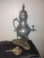 Front view of large antique Dutch pewter samovar with lid, three legs, spigot tap, and ornate trivet stand with floral detailing, sitting on black cloth background.