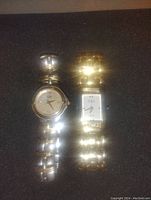 Two women's wristwatches placed side by side on a black surface. Left watch has a round face with silver-tone band; right watch has a rectangular face with gold-tone band.