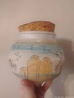 Front view of the ceramic jar showing hand-painted buildings, birds, and sky with large cork lid.