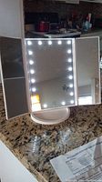 White tri-fold makeup mirror with LED circle lights on center panel, shown open and illuminated on counter.