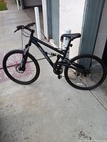 Right side view of black Gravity full suspension mountain bike leaning against a wall, showing frame, rear shock, drivetrain and wheels.