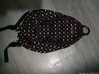 Photo of a black Dakine women's shoulder backpack adorned with white and pink geometric pattern, showing the front and adjustable shoulder straps, lying flat on a floor.