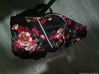 Photo of a black woman's shoulder backpack with bright floral print and a front zippered pocket, displayed on a wooden floor.