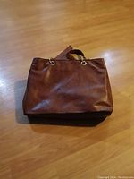 Brown leather Bueno women's purse laid flat on a wood floor, showing the front and main structure of the bag with gold-tone metal ring handle attachments.