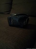 Black Guess makeup bag on brown fabric surface, showing embossed 'G' pattern and zippered top.