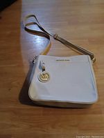 Michael Kors white leather crossbody purse pictured flat on wooden surface showing front side with logo charm and adjustable strap.