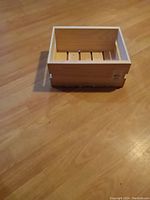 Small rectangular wooden box placed on a wood floor. Box has three wooden slats on the bottom and a smooth natural finish, showing slight marks of use.