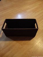 Rectangular dark brown wicker storage basket placed on wooden floor, showing open top and handle cutouts on shorter sides.