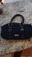 Front view of black Nine West purse with handles, buckle details, and brand plate on granite surface