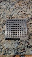 Square metal floor drain cover with grid pattern holes, two screws visible for mounting, placed on granite surface.