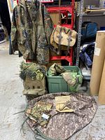 Overview of flecktarn jacket, blankets, canvas backpack with ghillie netting, loose netting on floor and Filson shoulder bag