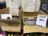 Open cardboard boxes showing boxed GreenPan cookware and Thermal Wall Cookware products inside, with labels visible from packaging and manuals.
