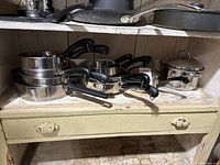 Shelved stack of various stainless steel pots and pans with black handles, showing assortment and count of items.