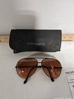 Front view of Serengeti aviator sunglasses with brown tinted lenses and black metal frame, placed below the black branded case on a white surface with measuring ruler in the background.