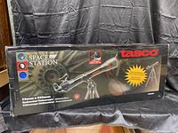 Box front view showing Tasco branding, Space Station name, and telescope image with tripod and red dot starpointer. Specifications: 70mm aperture, 800mm focal length, refractor telescope, altazimuth mount.