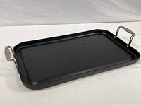 Black rectangular stove top griddle with raised stainless steel handles, viewed angled from side