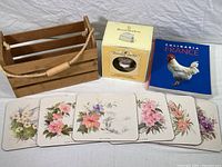 All items arranged showing wood storage box, cookbook, boxed teacup and saucer, and six small floral placemats with cork backing.