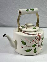 Side view of white enameled metal teakettle decorated with green holly leaves and red berries with red ribbon swirls, brass folding handle with decorative holly leaves on grip.
