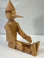 Full side view of the wooden Pinocchio sculpture sitting with legs extended and arms bent, showing articulated joints.