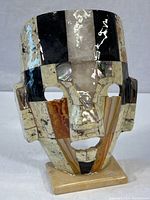 Front view of the stone mask sculpture showing the multi-colored mosaic tiles polished to a shine, mounted on a rectangular stone base.