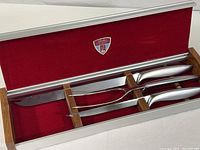 Photo of the aluminum storage box open, showing the carving set inside with three tools: carving knife, carving fork, sharpening steel with polished stainless handles.