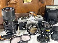 Photo showing Olympus OM-2 camera with 50mm lens attached, Vivitar 60-150mm lens, Vivitar 28mm lens, Olympus flash, lens caps, and vintage leather camera bag.