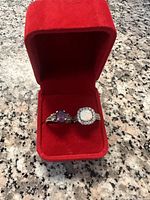 Two rings inside a red velvet ring box on a marble surface, one with a large opal stone and another with an amethyst stone.