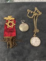 Shows the two pocket watches and the red ribbon medal with gold Star of David and crown insignia on black fabric background.