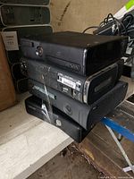 Stack of four black Xbox 360 consoles placed on surface, showing front and side views.