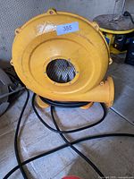 Yellow electric air blower with visible front mesh, coiled power cord, and model number sticker on the back side.