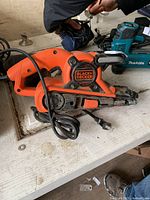 Black and Decker electric belt sander placed on a workbench alongside other power tools, showing detailed view of the sander with attached power cord and sanding belt area.