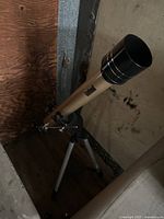 Side view of Bushnell telescope on tripod showing the full length of the silver tube and base stand. Located indoors against a wooden and metallic backdrop.