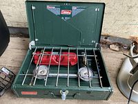 Green Coleman two-burner propane camping stove showing burners, metal grates, and red fuel tank.