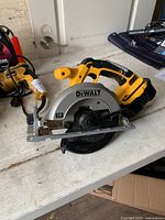 Photo shows a yellow and black DeWalt battery powered circular saw with an 18 gauge blade on a wooden table, alongside a yellow and black corded electric drill.