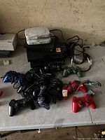 Two PlayStation 1 consoles stacked on top of one PlayStation 2 and two PlayStation 3 consoles with assorted controllers laid out in front, on a surface.