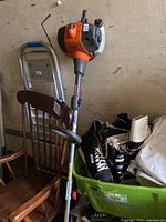 Photo of entire Husqvarna gas powered trimmer leaning against furniture (chair) and other stored items in background.