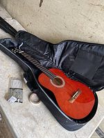 Red-orange Lucida acoustic guitar inside open soft black case with some repair materials nearby.