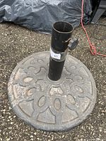 Photo showing a heavy round patio umbrella stand base with decorative cutout design, central black pole holder with adjustment knob, placed outdoors on gravel surface.