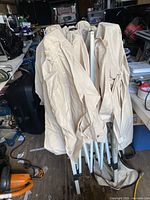 Photo showing the folded gazebo with beige fabric canopy attached to the white collapsible metal frame, partially folded and standing upright on a wooden floor in a storage environment.