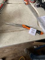 Image shows a small branch saw with an orange and black handle on a white wooden surface, accompanied by tags and other electrical equipment in the background.