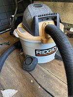 Ridgid wet/dry vacuum cleaner showing main body, handle, attached hose, and power cord with foot pedal on wooden surface.