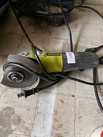 Yellow and black Ryobi corded angle grinder placed on tiled floor, cord visible, grinding disc attached, showing some dirt and wear from use.