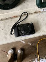 Black Sony Cyber-shot digital camera with wrist strap placed on a surface, showing lens and body details.